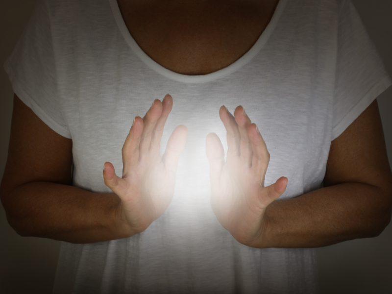 Woman standing in shadow with a white t shirt on, with her hands in front of her, with a light in between her hands. 