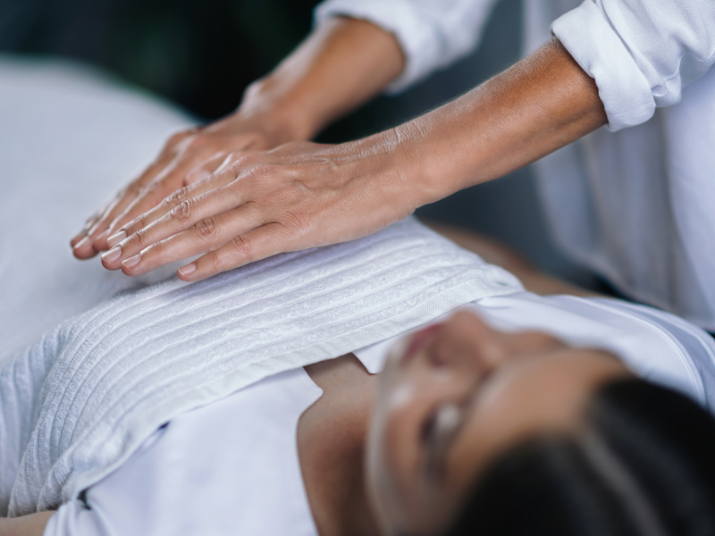 Woman on a treatment table, and a reiki practitioners hoovering over her heart chakra. White blanket covering the client.